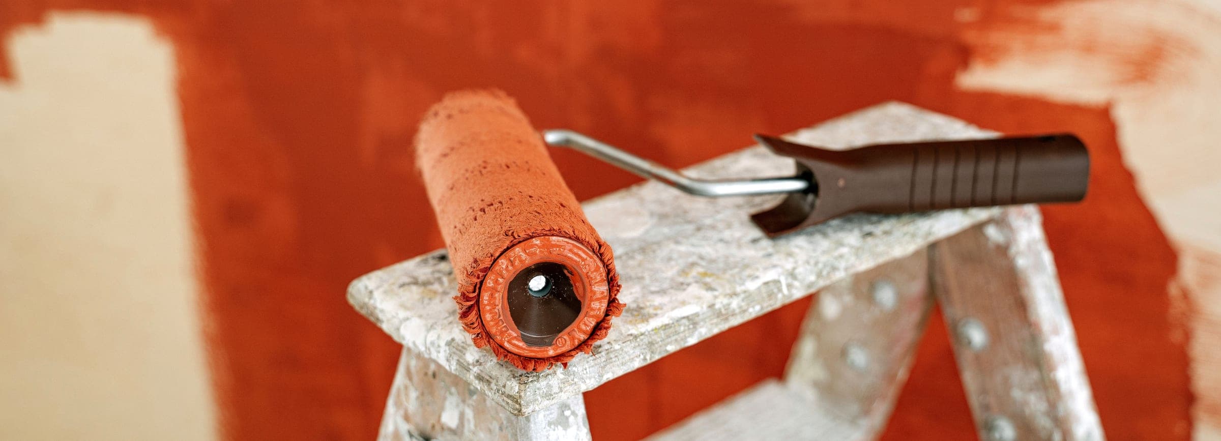 A terracotta-loaded paint roller resting on a splattered step ladder against a freshly painted wall