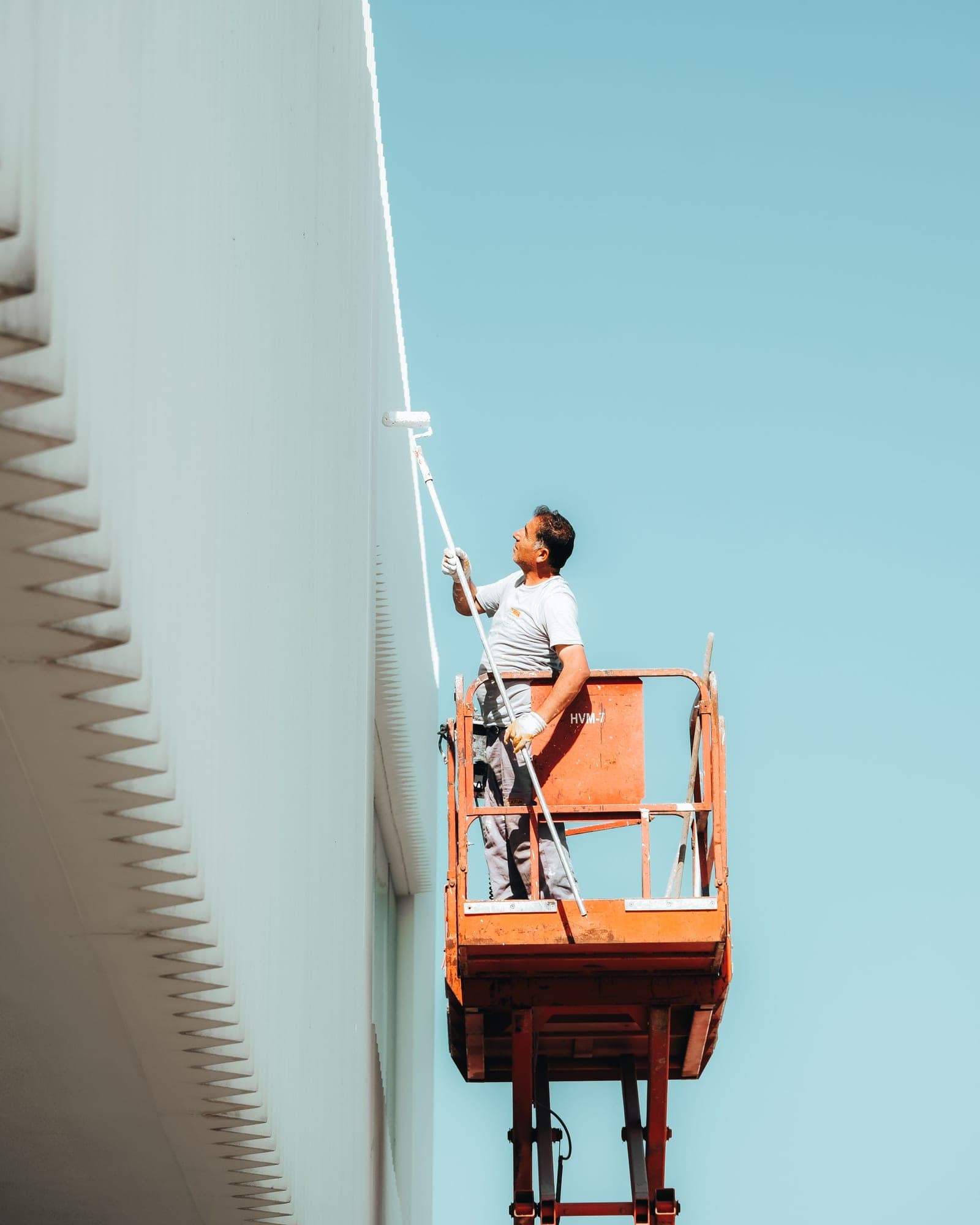 A painter on an orange lift rolling fresh white coating up a tall commercial wall under a clear sky